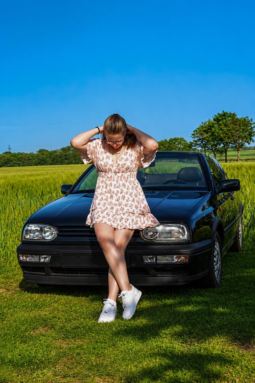 Frau im Blumenkleid vor einem schwarzen Cabrio an einem Feld